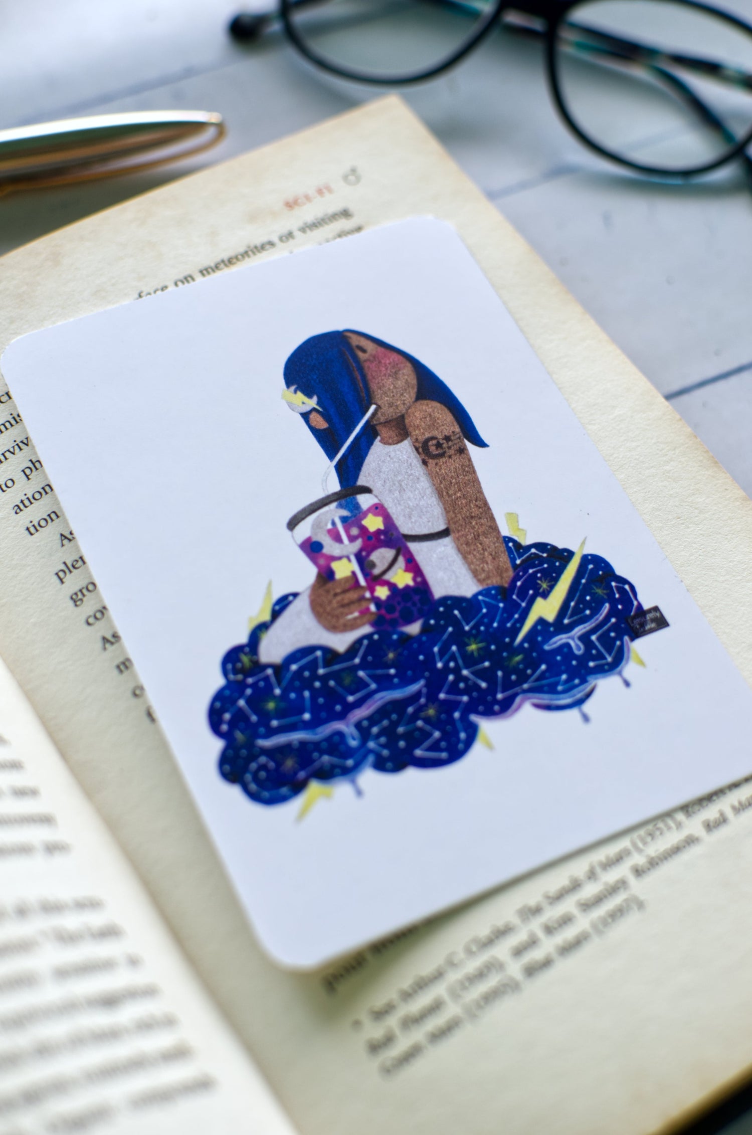 Hand-drawn art on bookmark, design is of a blue haired girl chilling on cloud sipping 'space juice', dreamy art